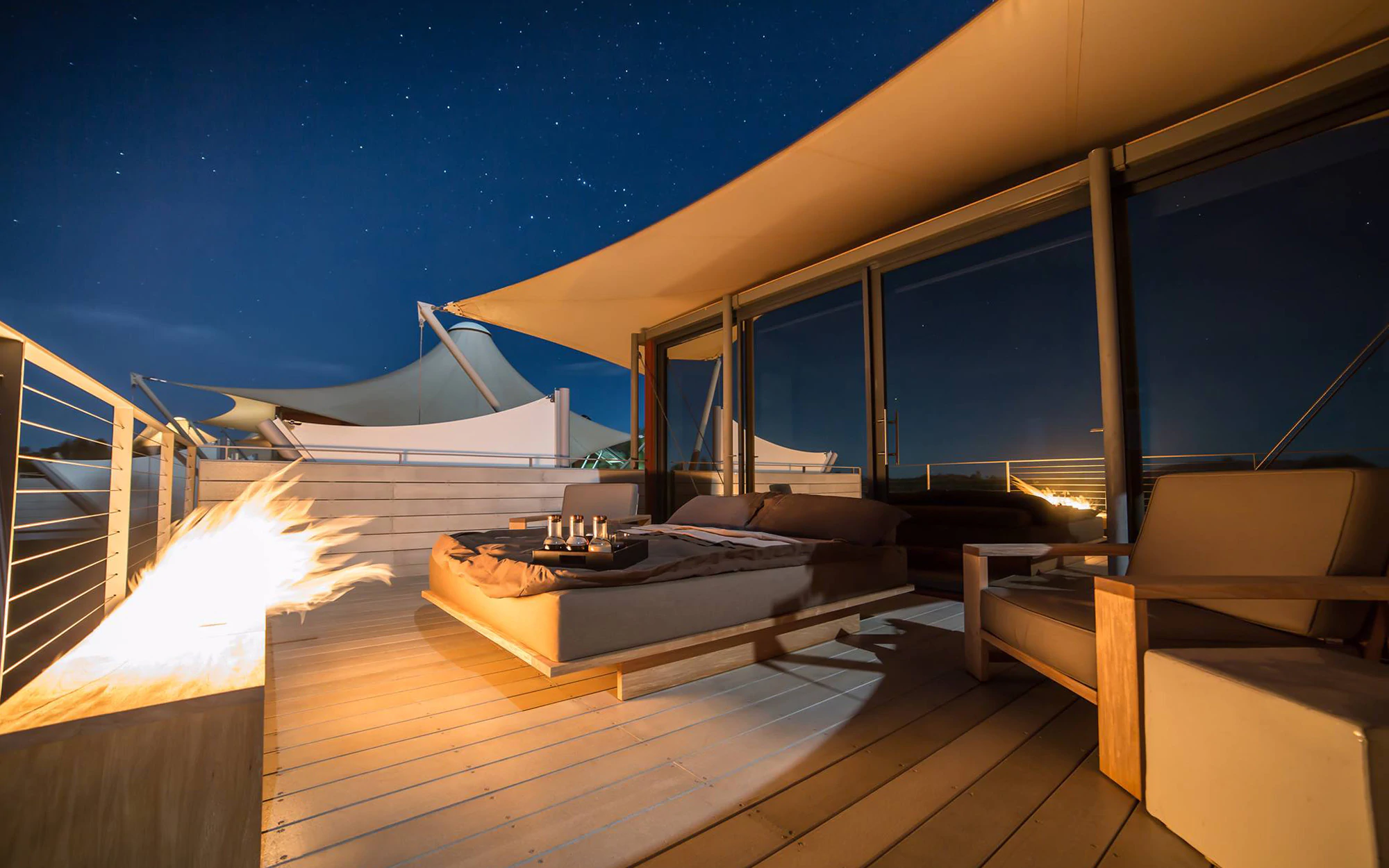 A bed for two people with a few glasses of champagne on a platform in the sea in front of a fireplace