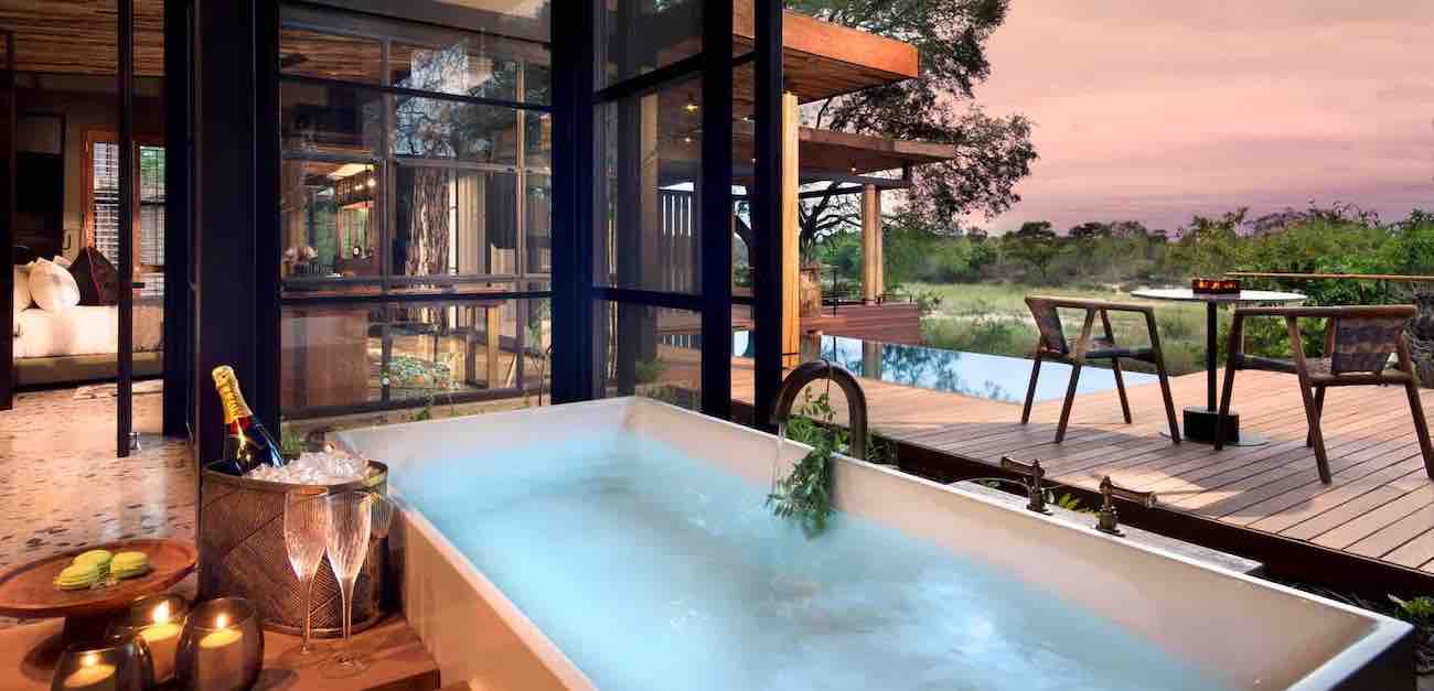 A jacuzzi with a bottle of champagne and two glasses with the view of a very modern style house and a wooden terrace with a table