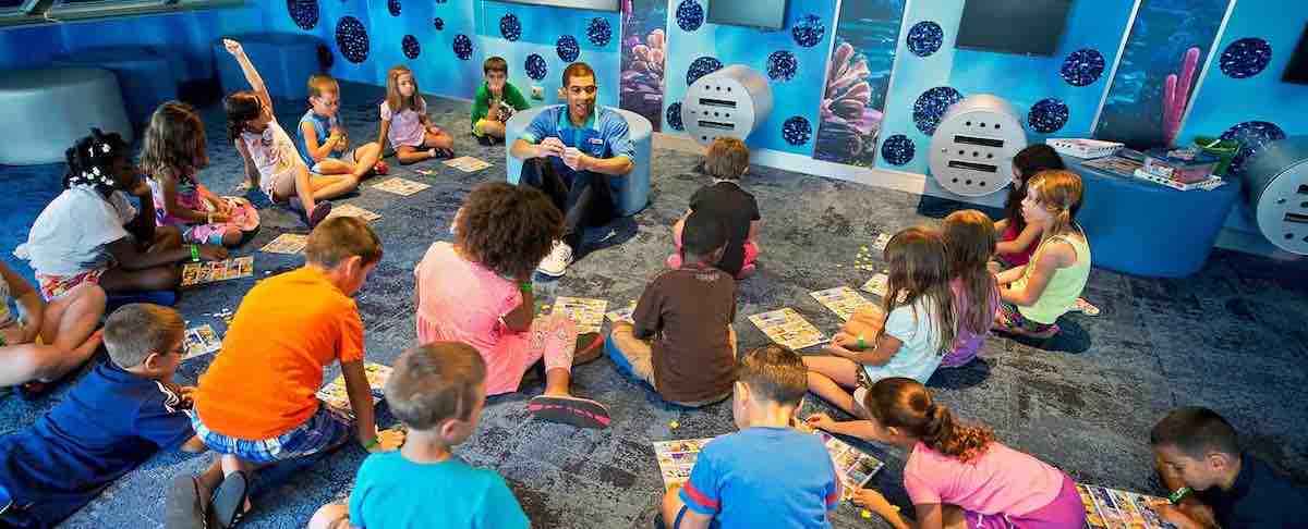 A group of children sitting on a carpet while a monitor explains a new game to them