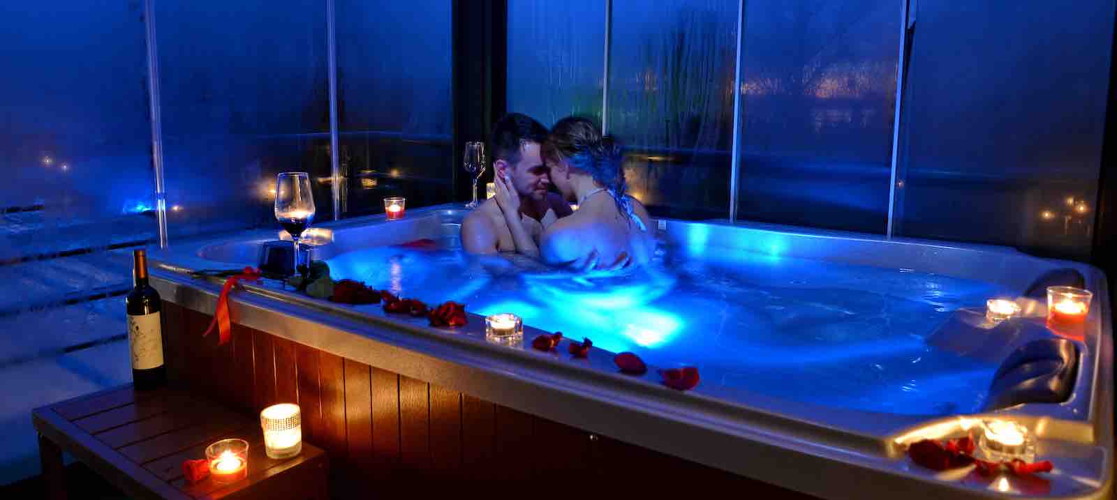 A couple embracing in a jacuzzi with some candles and a bottle of champagne with the lights off but with the chromotherapy lights on
